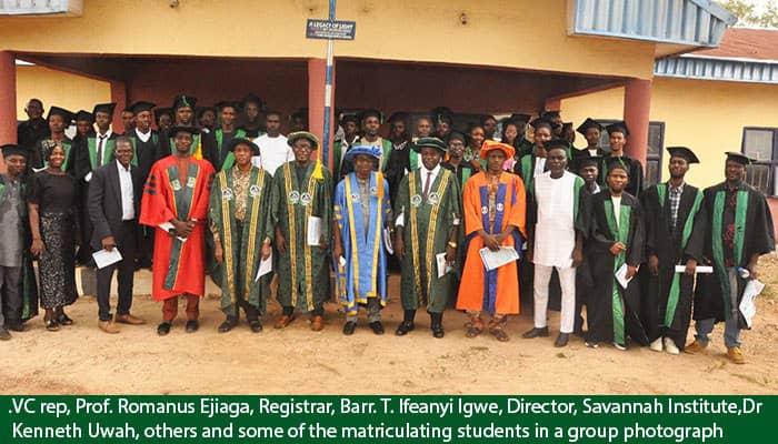 A group photograph of university officials and matriculating students in academic gowns posing in front of an academic building, including Prof. Romanus Ejiaga, Barr. T. Ifeanyi Igwe, and Dr. Kenneth Uwah.